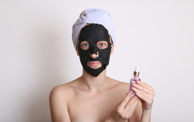 Beautiful woman with purifying black mask on her face and towel on head. Girl holding bottle of serum. White background. Facial skin care