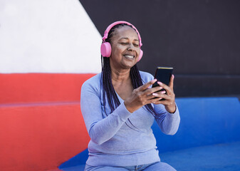 Happy senior african woman using mobile phone for listening music playlist with wireless headhone - Powered by Adobe