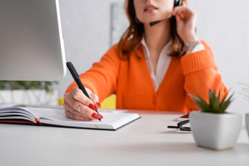 cropped view of blurred woman in headset writing in notebook while working at home