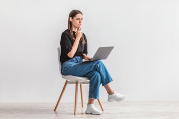 Thoughtful young woman using laptop while sitting on chair against white studio wall, full length