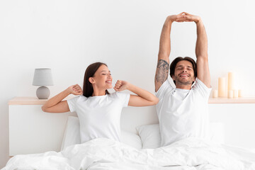 Happy caucasian millennial wife and husband woke up and stretching body sitting on bed in white bedroom