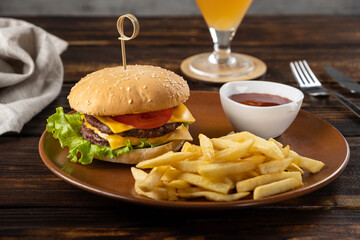 Homemade burger with juicy beef patty, tomatoes, cheese, sauce, lettuce leaves, French fries on a round plate on a dark wooden background. Fast food. Horizontal orientation, close-up, no people