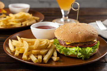 Homemade burger with juicy beef patty, tomatoes, cheese, sauce, lettuce leaves, French fries on a round plate on a dark wooden background. Fast food. Horizontal orientation, close-up, no people