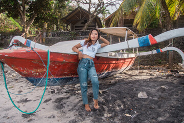 Young woman with red fishing boat dressed in blue jeans and white t-shirt posing on the beach. 