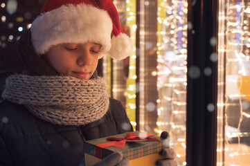 Boy in Santas hat with gifts box near illuminated shop window. Xmas presents holidays, or shopping on New Year or Christmas concept