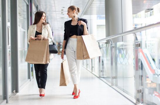 Two cheerful women friend with shopping bags talking while walking in the mall