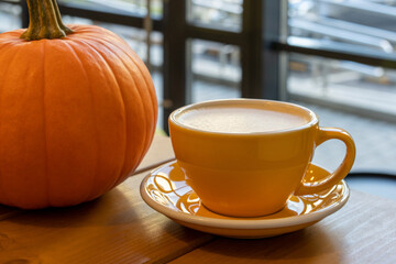 A cup of coffee in a yellow cup on a saucer and an orange pumpkin on a wooden table. Autumn. Cafe concept
