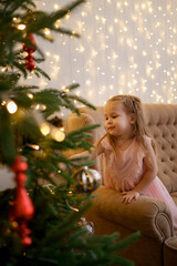 Little girl looking to a fir tree on Christmas tree making a wish while standing on sofa at home with lights in background