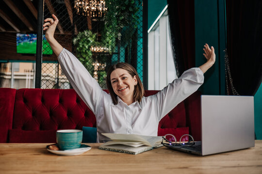 a happy brunette businesswoman in a white blouse is sitting at a table in a cafe with a phone and laptop