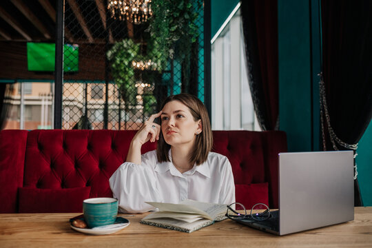 a thoughtful brunette businesswoman in a white blouse is sitting at a table in a cafe with a phone and laptop