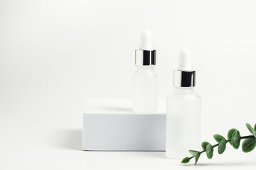 Two glass white cosmetic jars with pipettes dispensers. Eucalyptus branches. White square podium and background. Cosmetology and skin