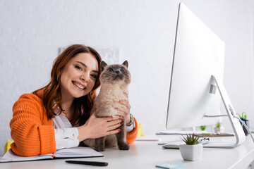 happy young freelancer looking at camera while hugging cat sitting on work desk