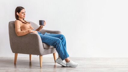 Full length of millennial woman drinking fresh coffee, having break in comfy armchair against white wall, panorama