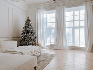 Studio decorations with stylish decorated Christmas tree with presents, toys, nutcracker next to big windows in white spacious living room with modern interior, sofa and gray carpet, Christmas morning