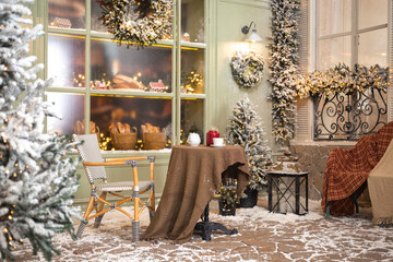 Christmas studio decorations outside in pastel green tones with a cafe table near bakery, door, wreaths, lanterns, garland, glass showcase with ginger bread, windows and many fir trees in snow 