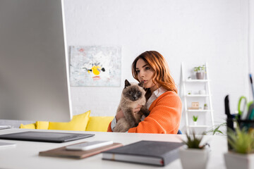 pensive freelancer sitting with cat at workplace and looking at monitor on blurred foreground