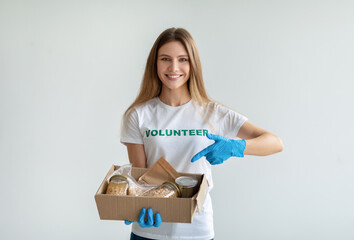 Happy female volunteer holding and pointing at box with donations, smiling at camera, standing over light background