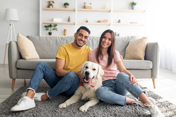 Full length portrait of positive international couple with their pet dog sitting on soft carpet at...