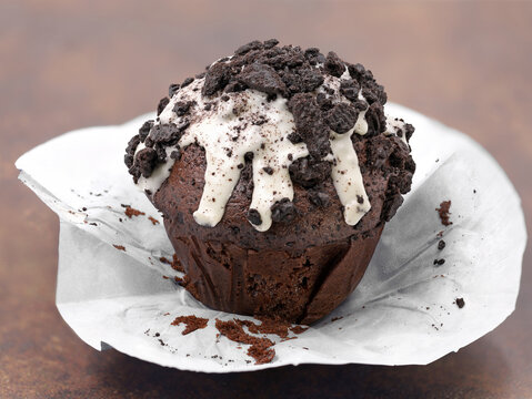 Chocolate Muffin With White Glaze And Dark Cookie Crumbs On Brown Background, Unpacked Cupcake