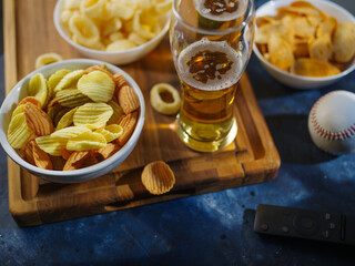 On a wooden tray, light beer in glasses, potato chips, onion rings. Nearby is a TV remote control and a ball. Watching your favorite sports programs, relaxing with friends.