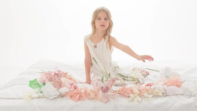 A little girl sits on a snow-white sheet among flowers on a white background.