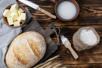 Fresh bread with a crispy crust on a dark brown wooden board. A mug of milk, butter in a clay bowl and a canvas bag with flour in the background. Top view