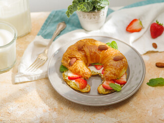 Appetizing composition. On a white plate, one ruddy croissant with butter, ripe fresh strawberries, fragrant mint leaves and crispy almonds. In the background is an indoor flower. Home comfort symbol.
