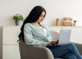 Focused Indian lady sitting in armchair with laptop pc, studying remotely or having online video conference at home