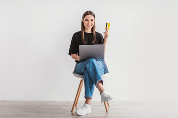 Beautiful young woman holding credit card and laptop, sitting on chair, ordering goods in web...