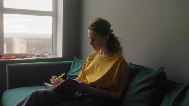 The girl makes notes in a notebook sitting on a bright sofa