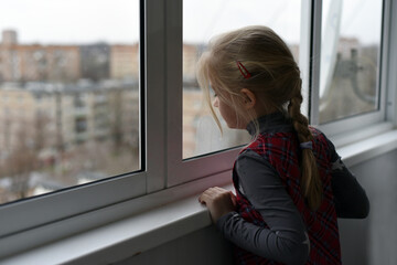 A girl with long white hair braided in a braid stands at the balcony window and looks at the city.