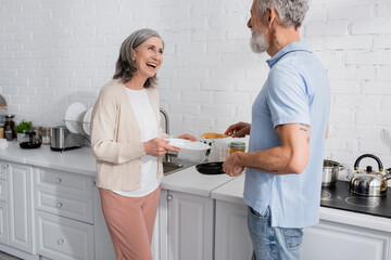 Positive woman holding plate near husband with frying pan and pancake in kitchen.