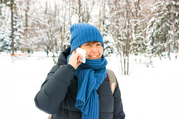 Fototapeta na wymiar A girl wearing blue scarf hat smiles talking on smartphone in snow-covered park woods in winter. Young European woman of 35-40 years old chatting cheerfully on phone standing in snow-covered landscape