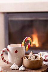 Large white mug with Christmas candy cane, sweets. Beautiful fireplace, flames on the background. Holiday atmosphere, magic, new year, warm cozy home, christmas concept. Nobody. Vertical. Copy space.