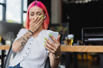 blurred businesswoman with pink hair and earphone using smartphone and covering mouth while laughing in office.