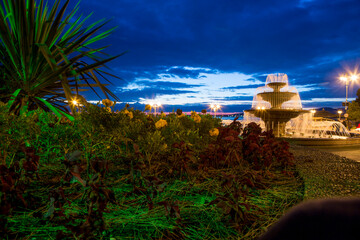 Singing three-tiered fountain and arch with columns. In the foreground are agave and rose bushes. Gelendzhik embankment in the evening. Lights are shining on the embankment with a balustrade.