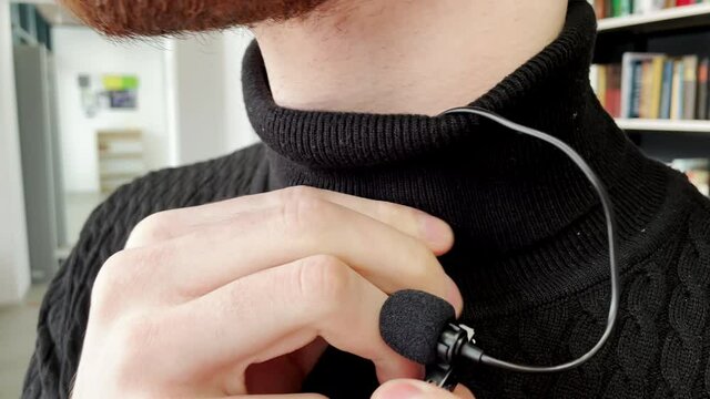 Young bearded man attaches small microphone to polo sweater collar against bookshelves in public library before interview closeup