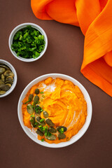 Pumpkin puree soup of butternut squash in bowl with green onions and pumpkin seeds, top view on plate with food on brown background