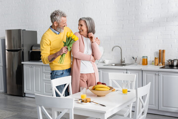 Cheerful man holding tulips near mature wife and tasty breakfast in kitchen.