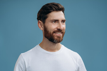 Fototapeta premium Portrait of young happy smiley man with white t-shirt looking away in front of the camera with smile. Studio shot, isolated on blue background