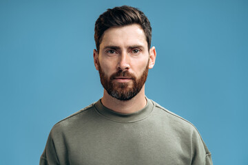 Portrait of attentive self confident bearded man looking at camera with serious expression, unsmiling determined business man. Indoor studio shot isolated on blue background