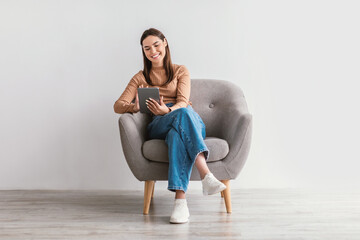 Positive young woman working online, sitting in armchair with digital tablet against white studio...
