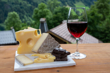 Cheese collection, Swiss cow cheese emmental, tomme and dark chocolate, glass of red wine from Savoie and mountains village in Apls on background