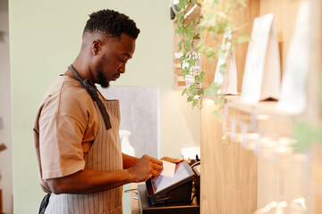 Fototapeta premium Horizontal side view shot of modern small cafe worker working with customer taking order using cash register