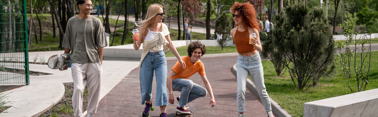 Fototapeta premium happy multiethnic friends skating in park with cocktails, banner.