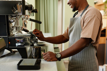 Fototapeta na wymiar Horizontal side view shot of young African American man working as barista in small cafe making espresso using coffee machine