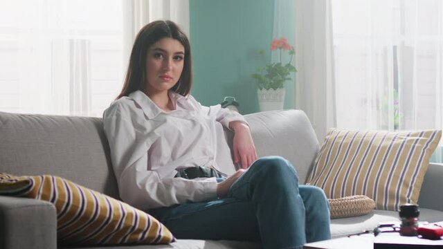 Portrait of young confident woman, dark-eyed brunette in white shirt and blue jeans, sitting on sofa and looking on camera, Slow motion.