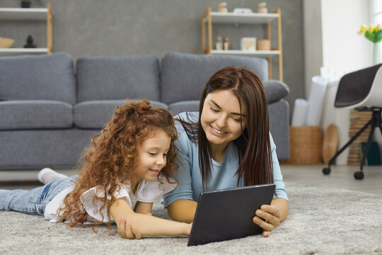 Modern technology. Young mother and her daughter use a digital tablet while lying together on the warm floor at home. Family surfing in the net while choosing movie or cartoons to watch at leisure.