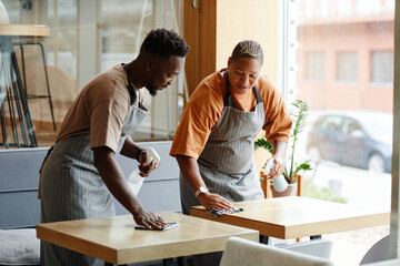 Young African American man and woman wearing aprons working in small cafe preparing tables for...
