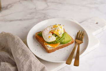 Savoury breakfast - A toast bread with cream cheese, avocado and poached egg on a white plate on marble tray